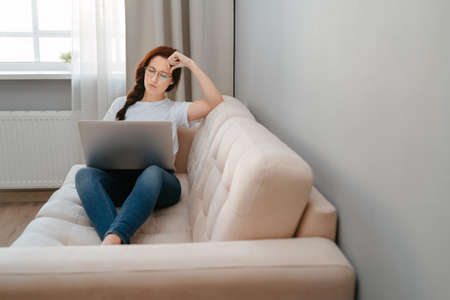 Young woman with glasses is sitting thoughtfully looking for goods and services online via a laptop.の写真素材