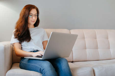A young woman works at home behind a laptop sitting on a beige couch. The concept of blogging and home learning.の写真素材