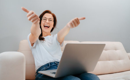 Smiling woman gives a thumbs-up to the camera while sitting with a computer on the couch at home. Concept of a satisfied user.の写真素材