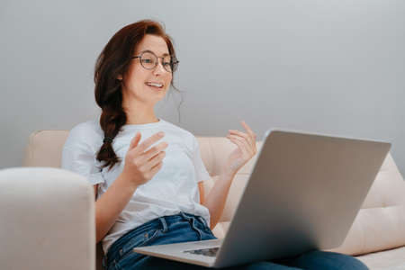 Young woman with glasses communicates on the Internet sitting on a bright sofa with a laptop at home. Concept of freelancing and learning home.の写真素材