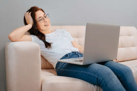 Young happy woman with glasses is resting at home on the couch ordering goods online on a laptop. Concept of purchases and sales via the Internet.の写真素材