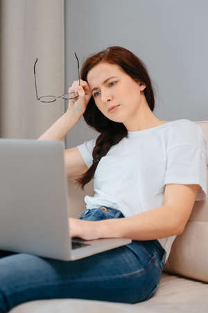 Young woman holding glasses in her hands reads messages on a laptop while sitting at home on the couch.の写真素材