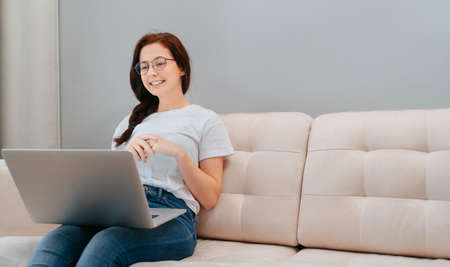 Young satisfied woman sitting on the sofa in the living room of the house listens to the interlocutor on the laptop online.の写真素材