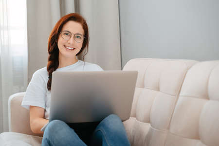Young girl learns at home on a laptop sitting on a beige couch. The concept blogging and online work.の写真素材