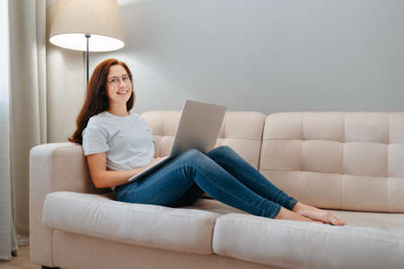 Young happy woman sits on a bright sofa at home with a laptop computer. Concept of online orders.の写真素材
