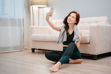 An athlete at home with a towel from sweat shoots a blog about her fitness achievements with her phone.の写真素材