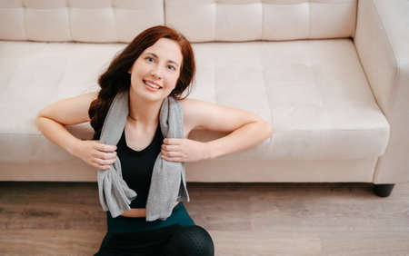 Female athlete is sitting on the background of a beige sofa with a towel from sweat after a yoga workout.Concept of a healthy lifestyle.の写真素材