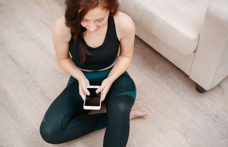 Athletic young woman using her mobile phone after an exercise session, sitting at home on the floor.Concept of home fitness.の写真素材