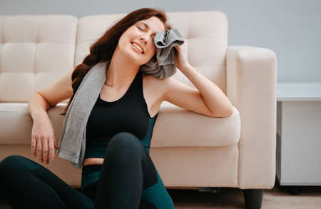 Young happy athlete wipes her sweat with a towel after doing sports at home. Concept of weight loss and fitness.の写真素材