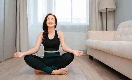 Sporty smiling woman is sitting in the lotus position doing stretching at home in a bright room.の写真素材
