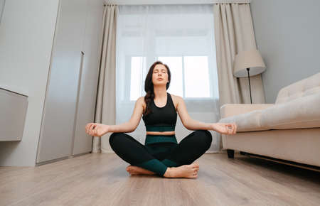 Sporty woman is sitting at home on the floor in the lotus position doing yoga. Concept of leisure at home.の写真素材