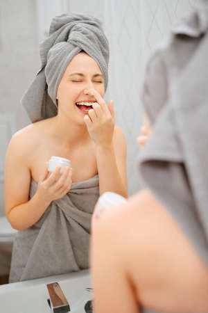 Cheerful woman in front of the bathroom mirror smears her face with a skin care cream. Concept of youth and cosmetology.の写真素材