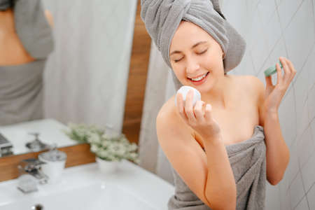 Happy young woman in a gray towel holds a package of face cream. Concept of care and youth.の写真素材