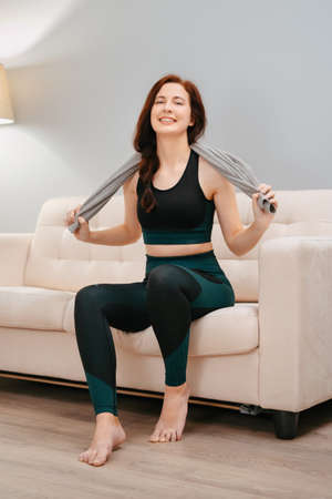Happy athlete is sitting on the sofa in the living room with a towel after intensive weight loss exercises.の写真素材