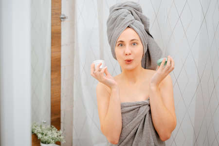 Young woman in a gray towel on the background of the bathroom holds a jar of serum for wrinkles and acne.の写真素材