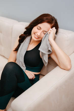 Female athlete is sitting on a beige sofa after a fitness workout wiping sweat with a towel.Concept of an active lifestyleの写真素材