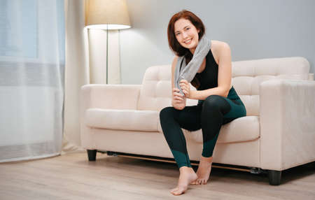 Female athlete is sitting on the couch resting after a fitness workout at home with a towel from sweat. Concept of weight loss.の写真素材