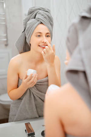 Young woman smears her face with white cream from wrinkles and acne in the bathroom looking in the mirror. Concept of cosmetology.の写真素材