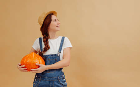 Young farmer woman in overalls holds a ripe orange pumpkin on a yellow background. Concept of organic food and halloween.の写真素材