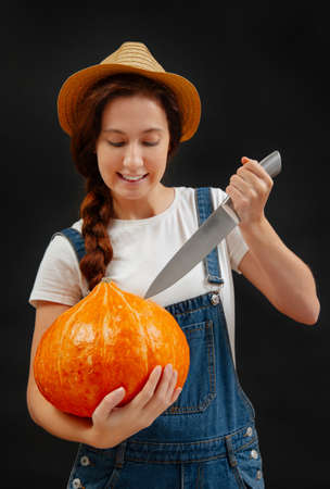 Woman in overalls on a black background cuts a lantern out of a pumpkin with a knife for the Halloween holiday.の写真素材