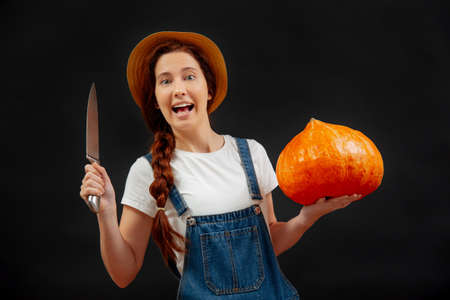 Farmer woman on a black background holds a pumpkin with a knife for carving a lantern for the Halloween holiday.の写真素材