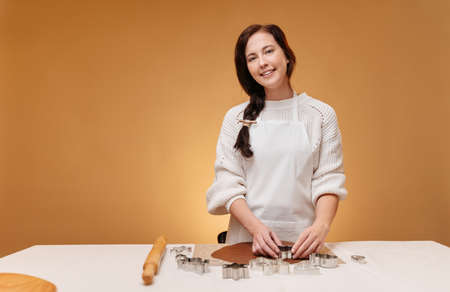 Young woman confectioner in a white apron prepares gingerbread cookies by cutting out metal forms on a yellow background. Concept baking new year.の写真素材