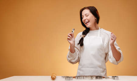 Joyful pastry chef woman in a white apron prepares gingerbread cookies for the holiday christmas on a yellow background.の写真素材