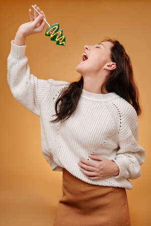 Young woman sings on a yellow background into a microphone of sweet lollipop in the form of a Christmas tree. New Year traditional holiday.の写真素材