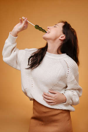 Young woman gnaws sweet caramel in the form of a Christmas tree on a yellow background.の写真素材