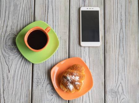 breakfast of coffee and a croissant on a wooden table, a mobile phone is next to each otherの写真素材