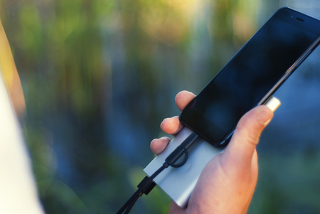 The guy is holding a portable charger with a smartphone in his hand. Man on a background of nature with a greenery and a lake.の写真素材