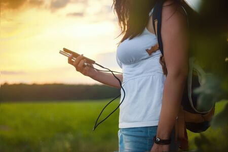 Girl with a backpack on a journey. Tourist holds in her hand a smartphone and a Power Bank on the background of the forest and sunset.の写真素材