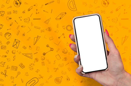 Back to School concept. Woman Hand holding the black Mockup smartphone with beautiful nails on the background with stationery icons. Mobile device isolated on orange colourの写真素材