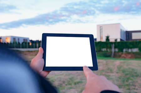 Mock up of a tablet with white screen in hand, against the background of modern housesの写真素材