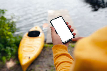 The tourist holds a phone in his hands. Mock up smartphone close-up on the background of a kayak and a lake. Concept on the topic of tourism and recreationの写真素材