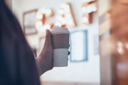 Cup of coffee in the hands of a guy in a cafe against the background of a luminous signboard. Mockup of a cardboard eco mugの写真素材