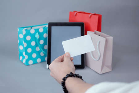 Online shopping concept. A girl holds a credit card in her hands, against the background of a tablet mockup with a white screen and beautiful gift bagsの写真素材