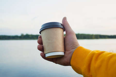 Man holds a cup of coffee in his hands on the background of nature with a lake.
