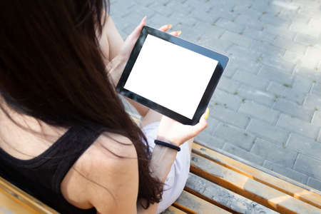 A girl sits on a bench and holds a tablet with a white screen in her hands. Mockup technology