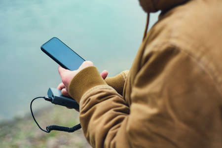 The guy holds a portable charger with a smartphone in his hand. A man on the background of a lake charges the telephone with Power Bank. Concept on the theme of tourismの写真素材