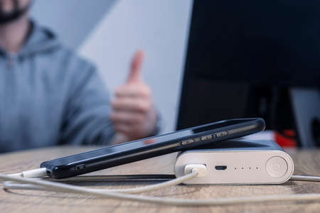Power bank with a phone and a cable on the background of a man sitting at the monitor. A portable charger charges a smartphone on a wooden tableの写真素材