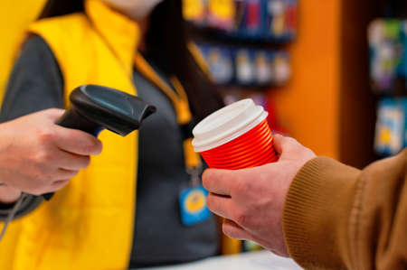 girl seller holds a barcode scanner in her hands and is counting on a man with a glass of coffee in...
