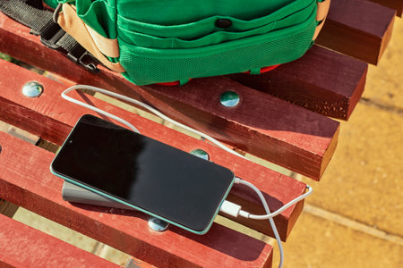 Powerbank charges a smartphone on a bench with a green bag. Portable charger for gadgetsの写真素材