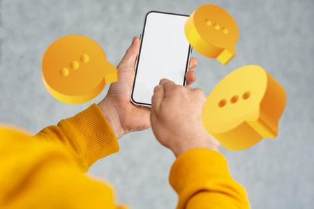 A close-up of a hand holding mockup smartphone with a blank screen, surrounded by floating yellow chat iconsの写真素材