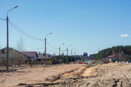 Construction of road and infrastructures with large sand piles sits near a forest and residential houses under a blue skyの写真素材