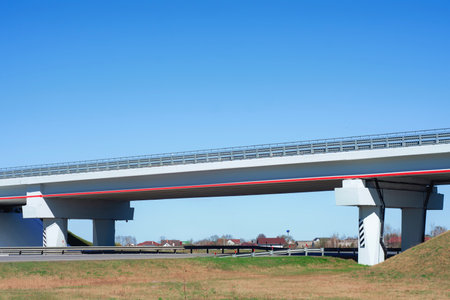 Highway overpass stretches across a clear blue sky, with a distant village visible in the...