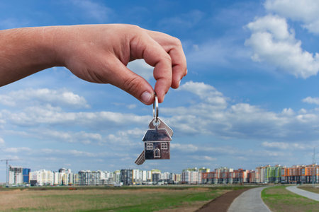 Man holds a house shaped keychain in the foreground, symbolizing real estate or new home ownership, with a modern cityscape and clear blue sky in the backgroundの写真素材