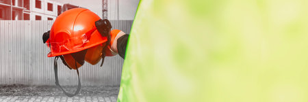 Worker in a high-visibility vest holds an orange hard hat with safety gear at a construction site,...