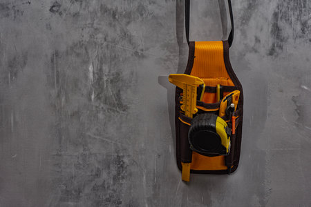 Tool belt with essential construction tools such as a measuring tape, utility cutter, pencil, and angle ruler hanging on a textured gray concrete wall. Tools for construction work, DIY and renovationの写真素材