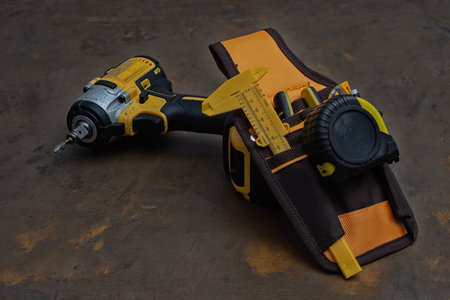 A tool belt holding a cordless drill, caliper, and tape measure lies on a worktable. Tools for...